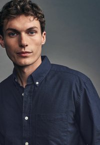 Námornícka košeľa s golierom, so skladovacou vreckom na prsiach a jemnou textúrou. Tkanina sa zdá byť ľahká a neformálna.