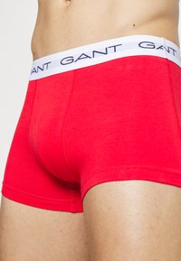 Rote Baumwoll-Boxershorts mit einem weißen Bund, der "GANT" in marineblauem Text zeigt. Glatte Textur mit einem schmalen Schnitt, der einen schlichten Stil betont.