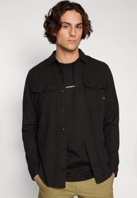 Μαύρο πουκάμισο με μακρύ μανίκι και σχέδιο με κουμπιά, δύο τσέπες στο στήθος και μαλακή υφή, που έχει τοποθετηθεί πάνω από ένα μαύρο μπλουζάκι.