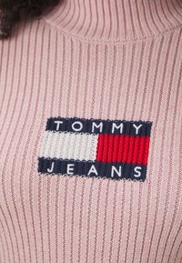 Maglione a coste rosa chiaro con un'etichetta tessuta "TOMMY JEANS" con dettagli blu navy, rossi e bianchi. Texture morbida e design con collo alto.