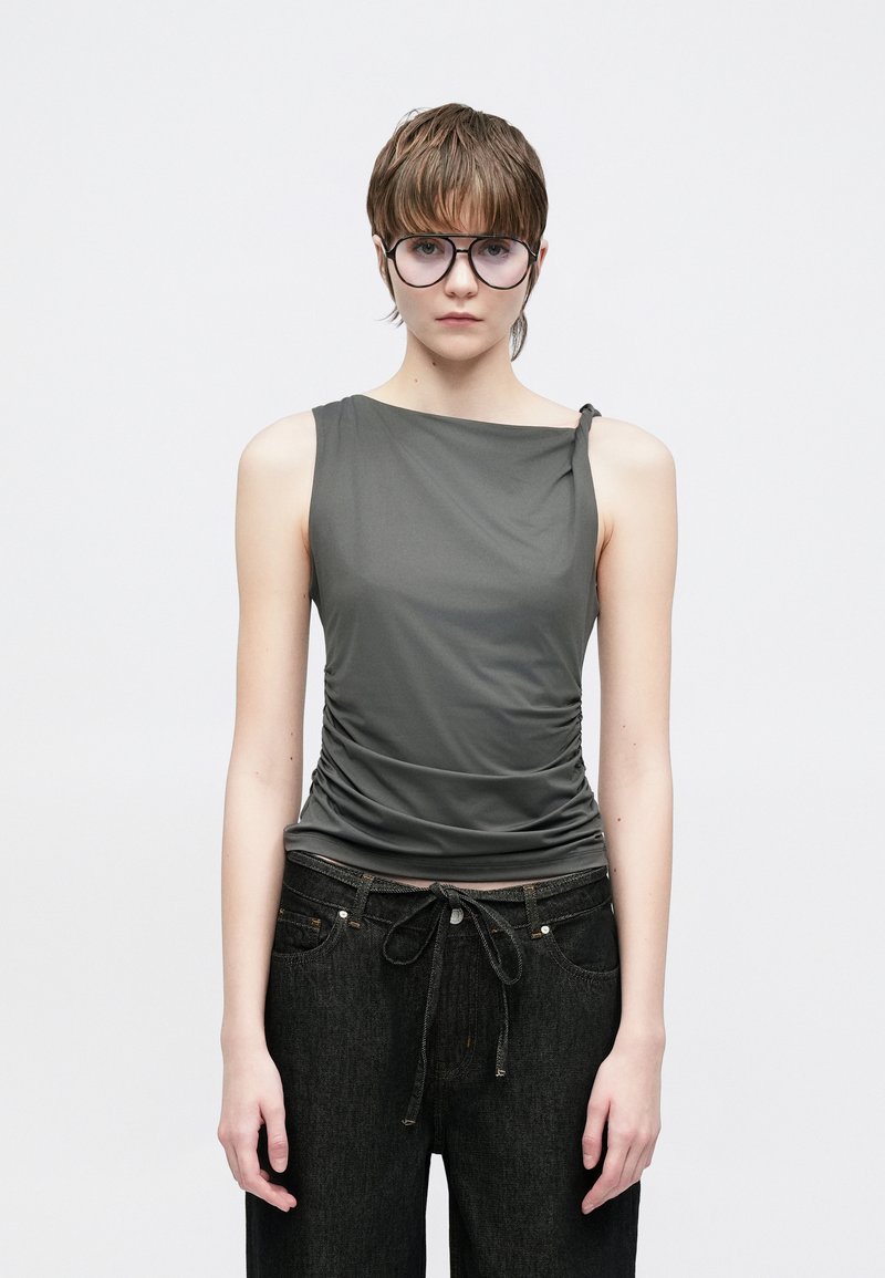 Γκρι αμάνικη μπλούζα με πιετά υφή και ασύμμετρο ντεκολτέ, συνδυασμένη με χαλαρά μαύρα τζιν παντελόνια με κορδόνι στη μέση.