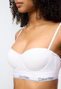 Γυναίκα που φοράει λευκό σουτιέν Calvin Klein με λεπτές τιράντες και ελαστική ταινία με λογότυπο, εμφανίζοντας τον λαιμό, τους ώμους και το πάνω μέρος του στήθους της.