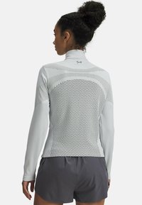 Helles graues, langärmliges Sportoberteil mit Mesh-Einsätzen am Rücken, hohem Kragen und tailliertem Schnitt, kombiniert mit dunklen Shorts.