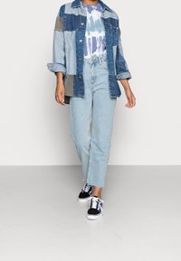 Chaqueta de mezclilla con apliques en azul oscuro y claro, llevada sobre una camiseta de tie-dye. Un pantalón vaquero azul claro y zapatillas bajas negras completan el atuendo.