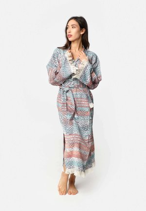 Vrouw staat op blote voeten, draagt een lange, met patronen bedrukte kimono met een ceintuur en franjes aan de randen, kijkt opzij tegen een effen achtergrond.