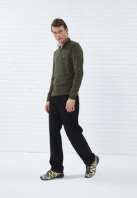 Pull olive avec col zippé, pantalon noir ample et baskets vertes avec des accents jaunes sur fond de briques blanches.