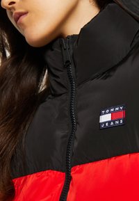 Čierna a červená zateplená bunda s vysokým golierom, vybavená zipsom a vyšitým logom "TOMMY JEANS" na hrudi.