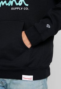 Handen i fickan på en svart hoodie med turkos "Diamond Supply Co." text, litet diamantlogo på ärmen och röd tagg vid fållen över ljusblå jeans.
