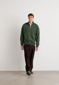 Maglione zip-up verde scuro con colletto alto, abbinato a pantaloni marroni e calzature verdi, su un pavimento grigio chiaro contro uno sfondo bianco.