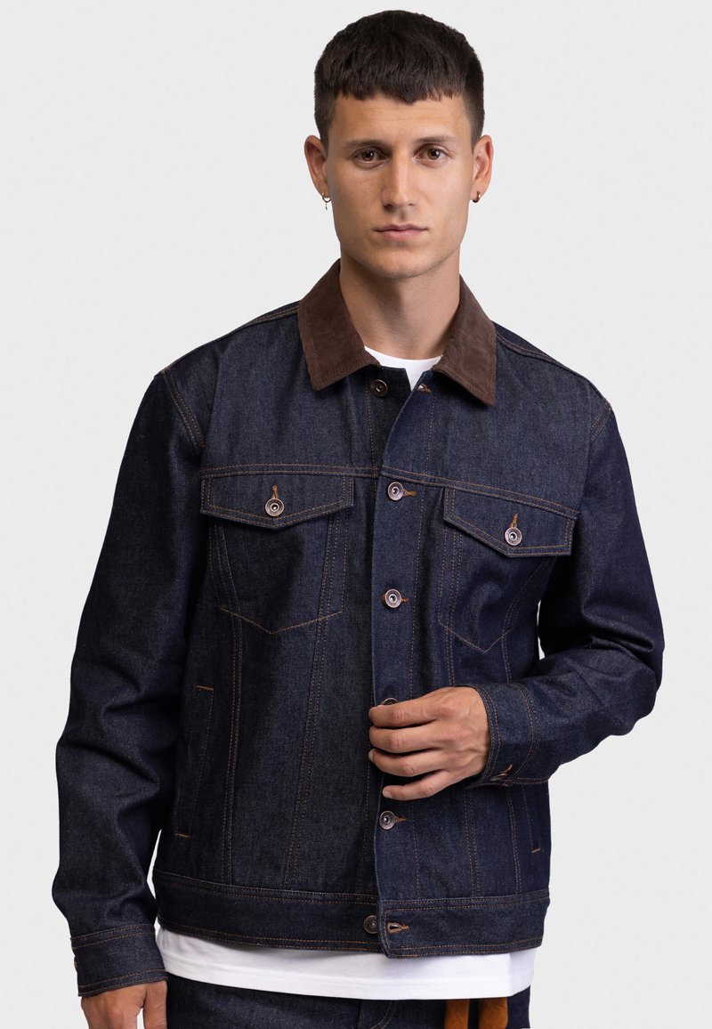 Νεαρός με κοντά μαλλιά φοράει σκούρο μπουφάν από τζιν με καφέ γιακά πάνω από λευκό πουκάμισο, στεκόμενος μπροστά σε απλό φωτεινό φόντο.