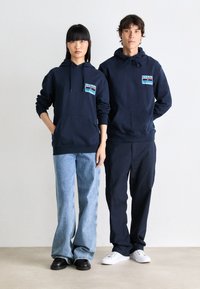 Sweatshirts marins avec poches avant, ornés d'un patch graphique coloré. Le modèle porte un jean évasé bleu clair et des mocassins noirs ; le deuxième modèle est en pantalon sombre et baskets blanches.