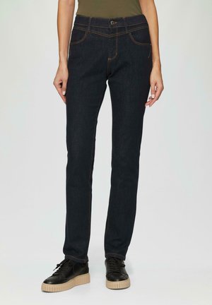 Jean en denim bleu foncé, avec une coupe slim, présentant des coutures orange, une fermeture à bouton et des poches avant. Porté avec des baskets plates noires.