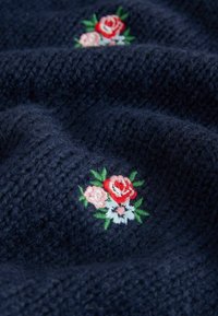 Námornícky pletený materiál s jemnou textúrou, zdobený vyšívanými ružami v červenej, ružovej a bielej farbe, obklopený zelenými listami.