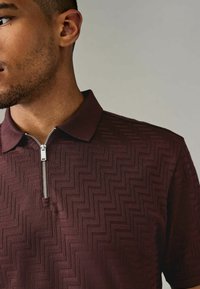 Polo burdeos con cuello con cremallera, que presenta un patrón de espiga texturizado. Manga corta y un corte relajado.