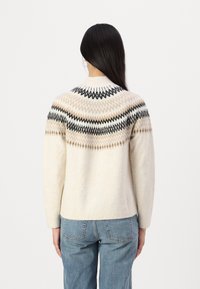 Cremefarbener Strickpullover mit einem gemusterten yoke in Schwarz und Beige, langen Ärmeln und rundem Halsausschnitt. Kombiniert mit blauen Jeans.