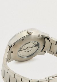 Vedere din spate a unui ceas de mână din metal argintiu cu mecanism intern mecanic vizibil și numele mărcii gravat pe spatele carcasei.