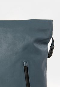 Blaue Lederhandtasche mit einem Reißverschlussfach, strukturiertem Oberflächen, gerolltem oberen Teil und verstellbarem Tragegurt mit einer Kunststoffschnalle und gewebten Details.