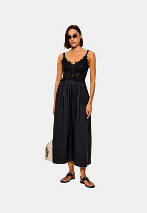 Donna che indossa un vestito nero senza maniche con corpetto in pizzo, sandali neri, occhiali da sole, che tiene una borsa bianca su uno sfondo semplice.