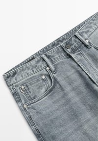 Jean en denim gris coupe droite, avec fermeture à boutons, rivets métalliques sur les poches, et une texture légèrement délavée.