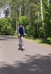 Cilvēks ar tumšu kreklu un ķiveri brauc ar melnu ceļa velosipēdu pa asfaltētu takas, ko ieskauj zaļa koku un augu vide.