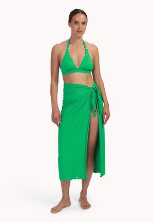 Kobieta stoi boso, ubrana w jaskrawozielony góra bikini na ramiączkach wiązanych za szyją oraz dopasowany sarong związywany w talii, z bocznym rozcięciem.