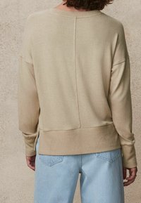 Pull à maille beige avec un bas et des poignets côtelés, présentant une couture centrale à l'arrière, associé à un jean bleu clair à coupe décontractée.