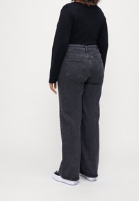 Černé tričko s dlouhým rukávem a tmavě šedé džínové kalhoty s vysokým pasem a širokými nohavicemi. Džíny mají zadní kapsy a strukturovanou látku. Viditelné bílé tenisky.