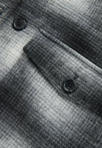 Grå og sort ternet stof med en struktureret overflade, med en enkelt sort knap på en klaffetaske.