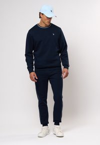 Jūrų laivyno treninginis kostiumas su baltais šonais, su maža logotipu ant krūtinės ir apvalia iškirpte. Šviesiai mėlyna kepurė.