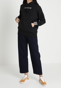 Mustat hupparit, joissa on valkoista brändäystä, yhdistettynä tummiin, väljiin farkkuihin ja vaaleisiin lenkkareihin, joissa on valkoisia yksityiskohtia.