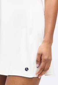 Κοντινό πλάνο ενός χεριού που ακουμπά δίπλα στο τελείωμα μιας λευκής φούστας με ένα μικρό μαύρο λογότυπο μπάλας του τένις κοντά στο κάτω άκρο.