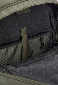 Intérieur d'un sac à dos en tissu vert avec une poche texturée, doublé de noir avec un motif répétitif "GUESS", et un curseur de fermeture éclair.