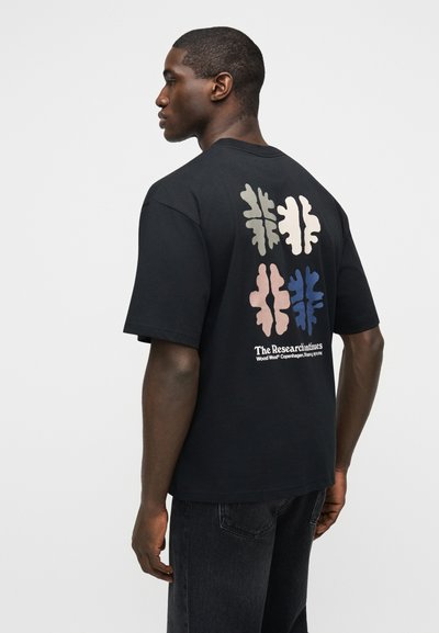 Άνδρας στραμμένος πλάγια φοράει μαύρο μπλουζάκι με τέσσερα αφηρημένα σχήματα φύλλων σε μπεζ, ροζ, γκρι και μπλε στο πίσω μέρος πάνω από λευκό κείμενο.