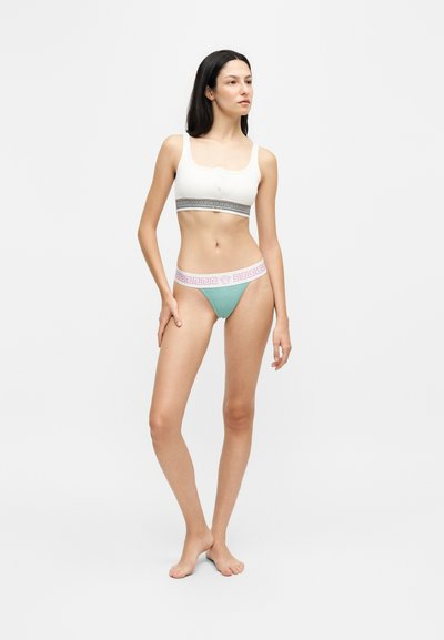 Femme debout pieds nus portant un crop top blanc et un sous-vêtement de style bikini vert avec des ceintures élastiques décorées sur un fond uni.