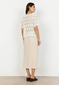 Haut en dentelle blanche à manches courtes avec un motif en zigzag, associé à une jupe midi beige et des mules en toile marron. Fente sur le côté de la jupe visible.