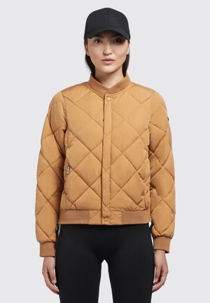 Veste bomber matelassée en marron, dotée d'une fermeture à pression, de poignets côtelés et d'une ceinture. Portée avec des leggings noirs et une casquette noire.