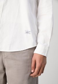 Camicia bianca a maniche lunghe realizzata in tessuto leggero, con orlo arrotondato e un piccolo etichetta del marchio sul lato, abbinata a pantaloni grigi chiaro.