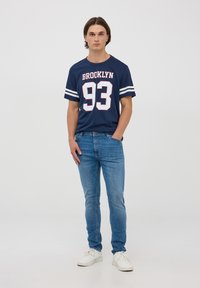 T-shirt in cotone navy con righe bianche e stampa "BROOKLYN 93", abbinata a jeans azzurri chiari e sneakers bianche, modello in posizione frontale.