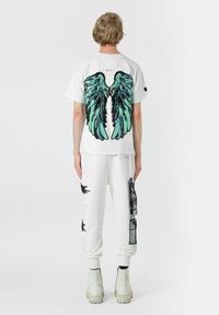 Camiseta blanca con un gran estampado de alas en negro y turquesa en la parte posterior, combinada con pantalones joggers blancos con gráficos en negro y un ajuste relajado.