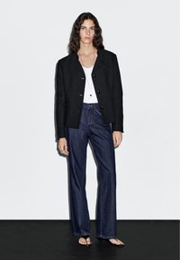 Žena v černé zapínané bundě, bílém topu, tmavě modrých širokých džínách, černých sandálech na podpatku a s vrstvami náhrdelníků stojící proti bílé zdi.