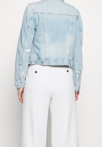 Personne portant une veste en denim délavé bleu clair avec des zips et un pantalon large blanc, montré de dos sur un fond uni.