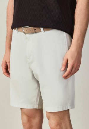 Mann in beigen Shorts mit braunem Ledergürtel und einem dunkel strukturierten Hemd, die Hände entspannt an den Seiten.