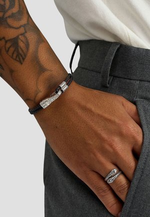 Black braided bracelet with a silver clasp featuring intricate designs. Model's hand displays a textured silver ring. Gray pants visible.