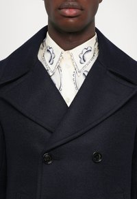 Detailný záber na tmavú osobu, ktorá má na sebe námornícky modrý dvojradový kabát cez smotanovú košeľu s čiernym kvetinovým vzorom na golieri, zapnutou až po krk.