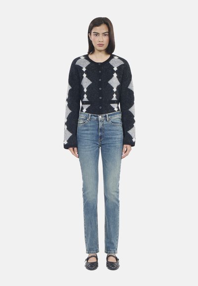 Cardigan in lana lavorato a maglia con motivo a diamante blu e bianco, bottoni neri, abbinato a jeans chiari a gamba dritta e scarpe nere.
