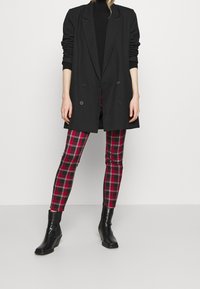 Blazer noir croisé à manches longues, associé à des leggings à carreaux rouges et noirs. Le mannequin porte des bottines noires. Texture de tissu lisse.
