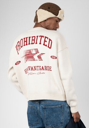 Uomo che indossa un maglione bianco con scritte rosse "PROHIBITED" e "BOVANTGARDE" sulla schiena, jeans blu e un cappello marrone foderato in pile.