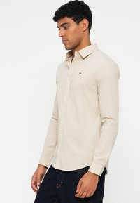 Lys beige knapp skjorte med spiss krage, lange ermer og en liten logo på brystet. Glatt, bomullslignende tekstur.