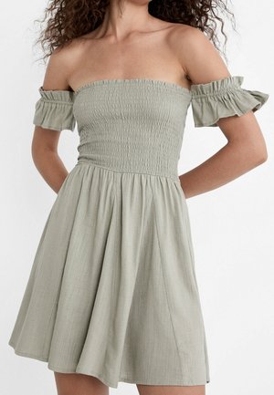 Femme portant une robe vert olive clair à épaules dénudées avec un corsage froncé et des manches à volants, debout devant un fond blanc.