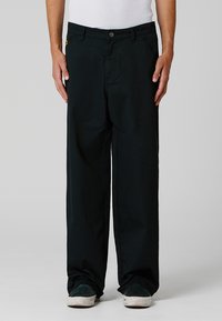 Zwarte wijde broek gemaakt van een gladde stof. Beschikt over een knoopsluiting, riemlussen en voorzakken. G gedragen met casual witte schoenen.